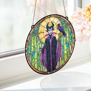 Puede incluir: Arte de ventana de vidriera ovalada que presenta una ilustraci&oacute;n estilizada de un personaje con cuernos negros, una capa morada y negra y un cuervo en un bast&oacute;n. La obra de arte tiene un borde negro y est&aacute; suspendida por una cadena.