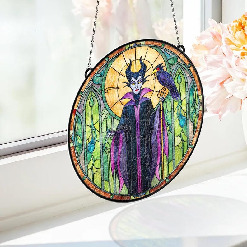 Puede incluir: Arte de ventana de vidriera ovalada que presenta una ilustraci&oacute;n estilizada de un personaje con cuernos negros, una capa morada y negra y un cuervo en un bast&oacute;n. La obra de arte tiene un borde negro y est&aacute; suspendida por una cadena.