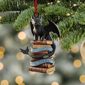 May include: A black dragon ornament perched atop a stack of books, with a blue banner that reads "in a world of bookworms" and "be a book dragon". The ornament is hanging from a red string, with a blurred background of a Christmas tree.