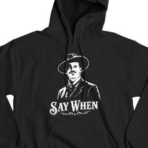 May include: Black hoodie featuring a white graphic of a man in a hat and suit, with the text "SAY WHEN" in a stylized font. The design is centered on the front of the hooded sweatshirt.
