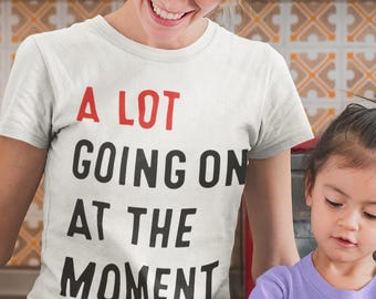 May include: A white t-shirt with the text "A LOT GOING ON AT THE MOMENT" in red and black. A woman and a child are in a kitchen, with ingredients and cooking utensils on a wooden table. A red microwave is in the background.