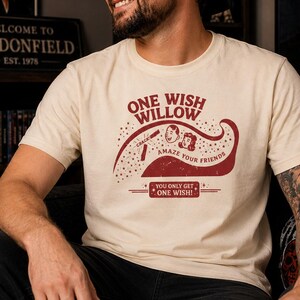 May include: A cream-colored short-sleeve t-shirt featuring a maroon graphic print. The design includes the text 'ONE WISH WILLOW', 'AMAZE YOUR FRIENDS', and 'YOU ONLY GET ONE WISH!', alongside a stylized illustration of two faces and a magic wand.