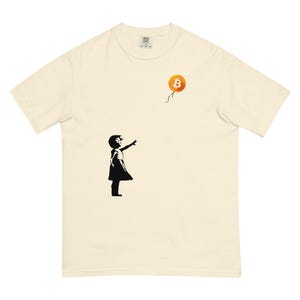 May include: Cream-colored t-shirt featuring a black silhouette of a girl reaching for a Bitcoin balloon. The Bitcoin symbol is orange and the balloon string is black. The shirt is a comfortable, casual style.