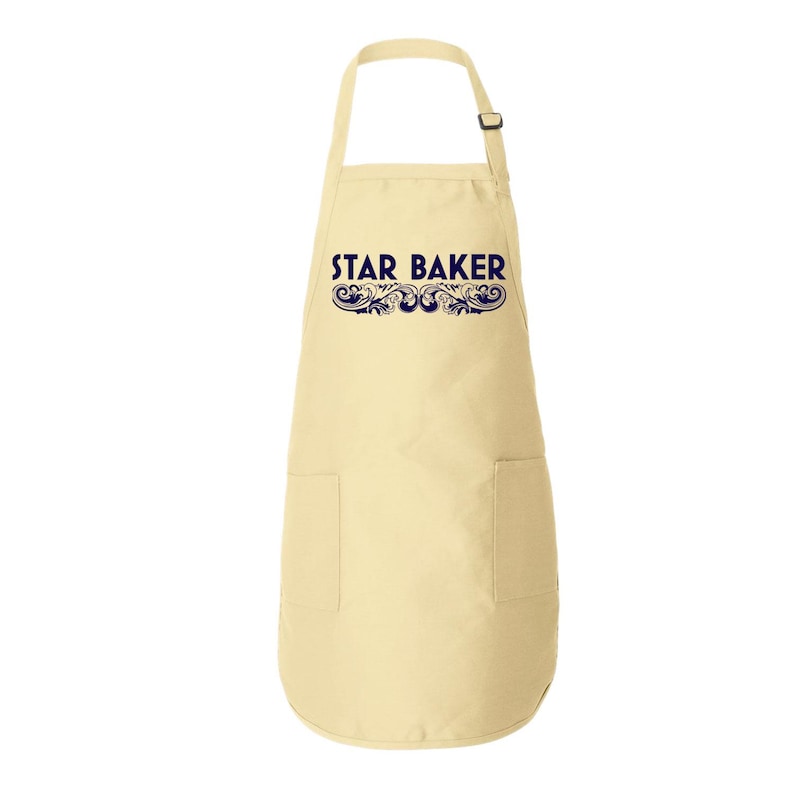 Op de afbeelding: Een lichtbeige schort met de tekst STAR BAKER in donkerblauwe letters, vergezeld van een decoratief donkerblauw krulpatroon eronder. Het schort heeft een verstelbare nekband met een zwarte gesp en twee voorzakken.
