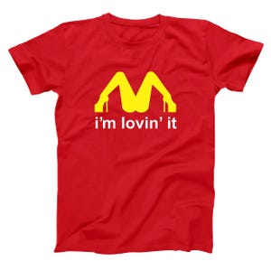 May include: Red t-shirt with a yellow graphic of legs in high heels forming an "M" shape, with the phrase "i'm lovin' it" printed below in white. The shirt is a casual wear item.