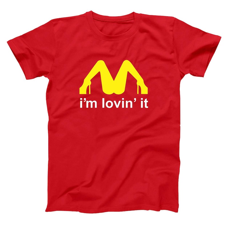 May include: Red t-shirt with a yellow graphic of legs in high heels forming an "M" shape, with the phrase "i'm lovin' it" printed below in white. The shirt is a casual wear item.