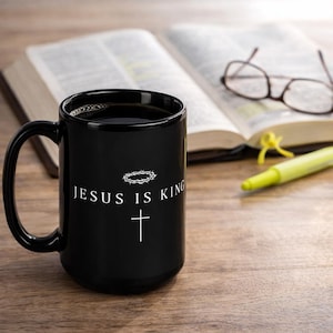 May include: A black ceramic mug with the text "JESUS IS KING" and a cross graphic. The mug is filled with a dark liquid and sits on a wooden surface next to an open book, glasses, and a highlighter.