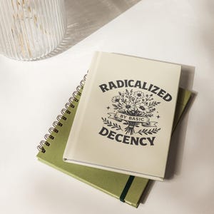 May include: Two notebooks, one spiral-bound and the other with a hard cover, are stacked on a white surface. The top notebook has the text "RADICALIZED BY BASIC DECENCY" with a floral design. The bottom notebook is olive green.