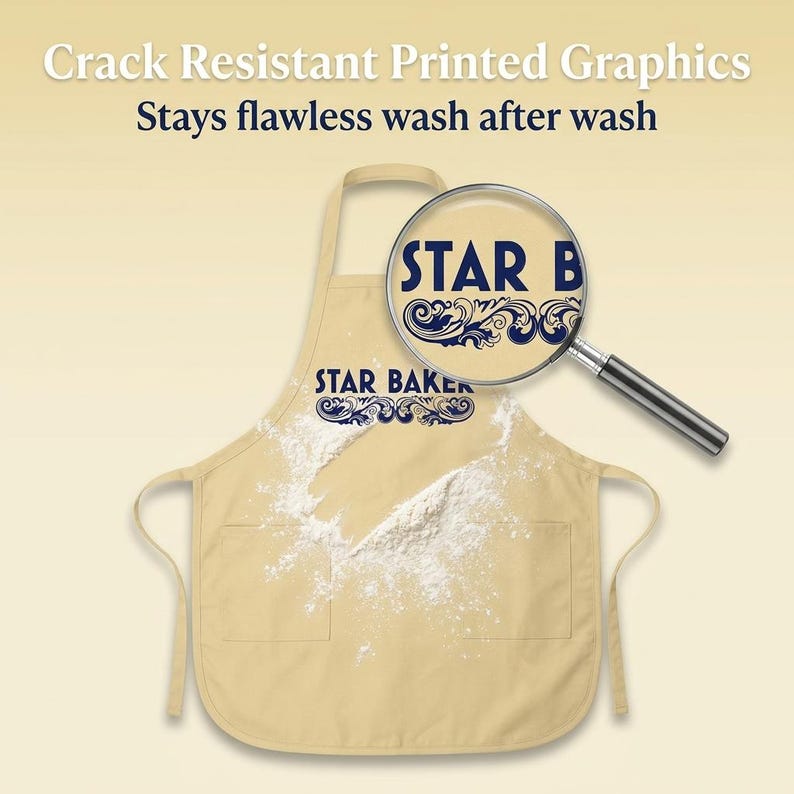 Op de afbeelding: Een beige schort met de tekst STAR BAKER in donkerblauw met decoratieve krullen. Een vergrootglas benadrukt de afdrukkwaliteit. Witte bloem is over de voorkant gestrooid. De tekst luidt Crack Resistant Printed Graphics.
