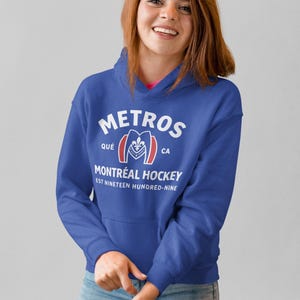 May include: A royal blue hooded sweatshirt featuring white text that reads METROS, QU&Eacute; CA, MONTR&Eacute;AL HOCKEY, and EST NINETEEN HUNDRED-NINE, with a central graphic of a stylized letter M containing a fleur-de-lis and the year 1909 in red and white.
