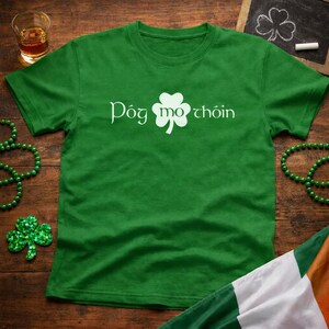 May include: A green t-shirt with the text "Póg mo chóin" in white, along with a white shamrock. The shirt is displayed on a wooden surface, with a glass of amber liquid and green beads nearby. An Irish flag is in the corner.
