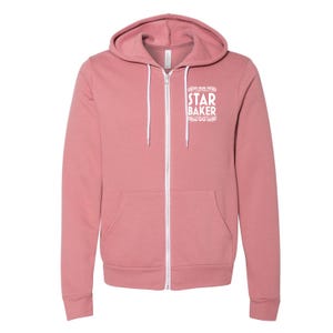 May include: A dusty rose zip-up hoodie with a white zipper and drawstring. The hoodie features a front pocket and the words "STAR BAKER" in white script on the upper chest. The hoodie is made of a soft material.