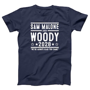 May include: Navy blue t-shirt with white text that reads "SAM MALONE AND WOODY 2028" with stars and the phrase "WE'RE ALWAYS GLAD YOU CAME". A casual tee for fans of a TV show.