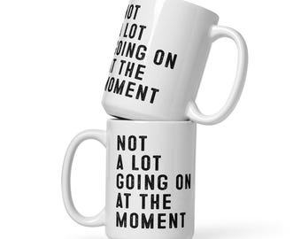 May include: Two white ceramic mugs stacked on top of each other. Each mug has a large handle and the words "NOT A LOT GOING ON AT THE MOMENT" printed in black, centered on the mug.