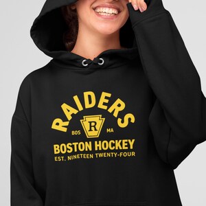 May include: A black hooded sweatshirt featuring yellow screen-printed text that reads RAIDERS, BOSTON HOCKEY, and EST. NINETEEN TWENTY-FOUR, with a central yellow emblem containing the letter R and the letters BOS and MA on either side.