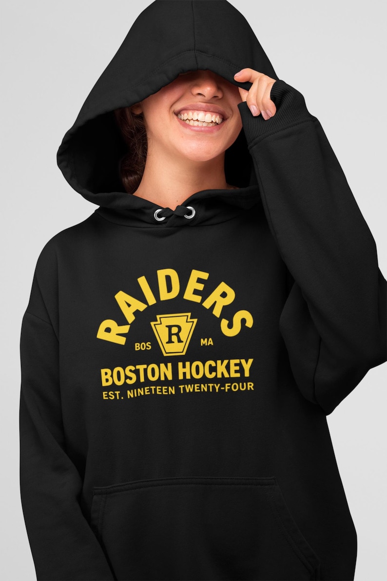May include: A black hooded sweatshirt featuring yellow screen-printed text that reads RAIDERS, BOSTON HOCKEY, and EST. NINETEEN TWENTY-FOUR, with a central yellow emblem containing the letter R and the letters BOS and MA on either side.