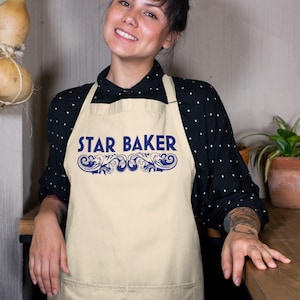 Op de afbeelding: Een beige schort met de tekst STAR BAKER in blauwe hoofdletters, geplaatst boven een blauw decoratief krulontwerp. Het schort heeft een grote voorzak en wordt gedragen over een zwart shirt met lange mouwen en witte stippen.