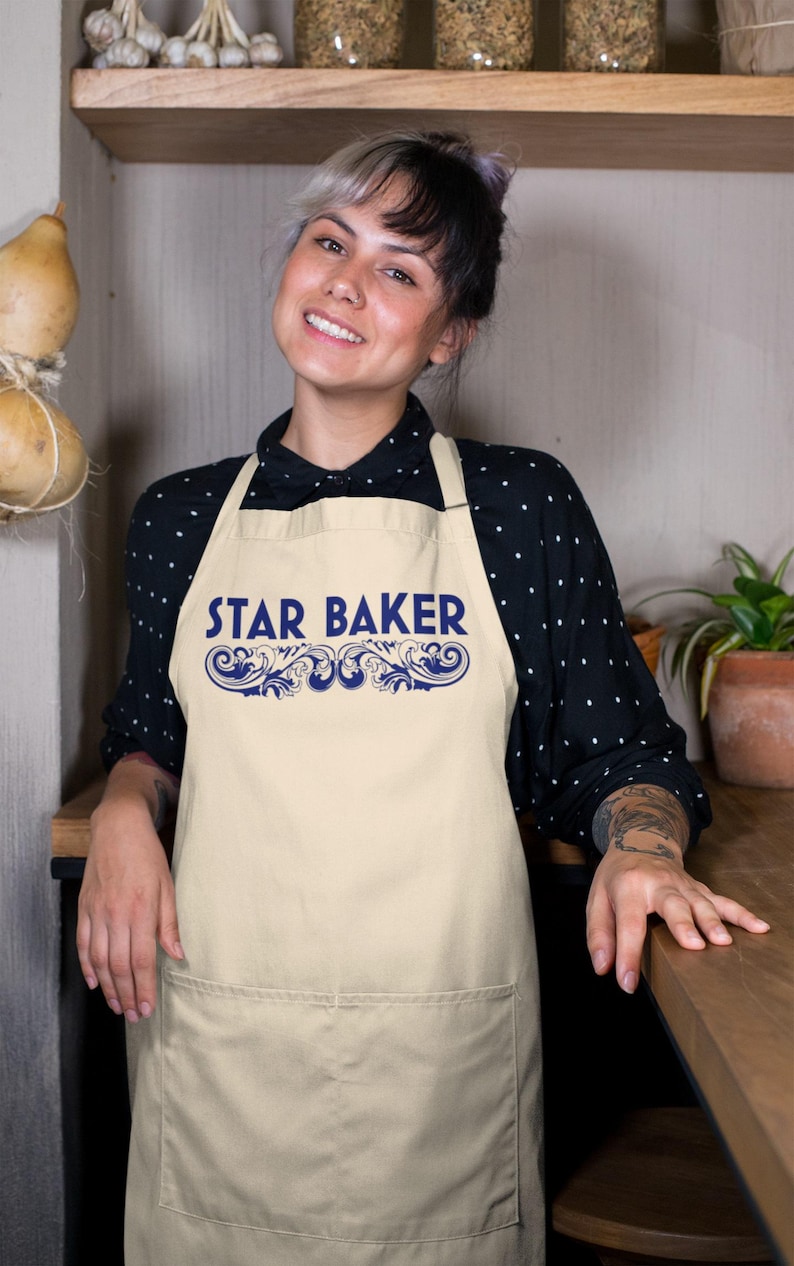 Op de afbeelding: Een beige schort met de tekst STAR BAKER in blauwe hoofdletters, geplaatst boven een blauw decoratief krulontwerp. Het schort heeft een grote voorzak en wordt gedragen over een zwart shirt met lange mouwen en witte stippen.