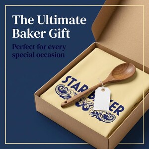 Op de afbeelding: Een lichtgeel stoffen item met donkerblauwe tekst STAR BAKER en decoratieve krullen, geplaatst in een kartonnen doos. Een houten lepel met een leeg wit rechthoekig label bevestigd met een touwtje ligt bovenop de stof. De doos staat open.