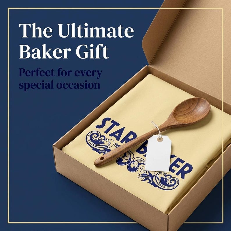 Op de afbeelding: Een lichtgeel stoffen item met donkerblauwe tekst STAR BAKER en decoratieve krullen, geplaatst in een kartonnen doos. Een houten lepel met een leeg wit rechthoekig label bevestigd met een touwtje ligt bovenop de stof. De doos staat open.