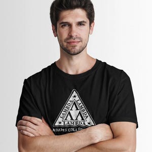 Pu&ograve; includere: T-shirt nero girocollo con un design triangolare bianco invecchiato. Il design include la parola "LAMBDA" e il testo "ADAMS COLLEGE". La persona indossa la maglietta con le braccia incrociate.