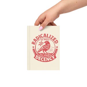 May include: A cream-coloured journal featuring a red graphic with the text "RADICALIZED BY BASIC DECENCY." The graphic includes a bird with a branch. The journal is held by a hand.