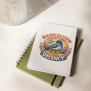 May include: A white journal with the text "RADICALIZED BY BASIC DECENCY" and a bird illustration, on top of a green spiral notebook and a green notebook with an elastic band. A clear glass vase is in the background.
