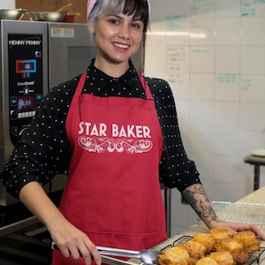 May include: A red apron with the text "STAR BAKER" in white, worn by a person in a kitchen. The person is holding tongs over a tray of golden-brown pastries. A Henny Penny oven is visible in the background.