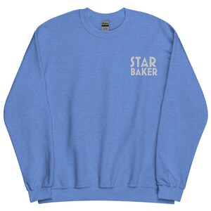 May include: A light blue sweatshirt with the text "STAR BAKER" in a light grey color. The sweatshirt is a crew neck style and appears to be made of a soft material. The text is centered on the upper chest area.