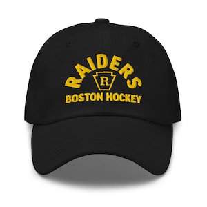 May include: Black baseball cap with the word "RAIDERS" arched across the front in yellow, above "BOSTON HOCKEY". The word "RAIDERS" has a yellow emblem in the center with the letter "R".
