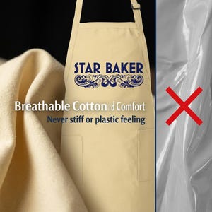 Op de afbeelding: Een beige schort met de tekst STAR BAKER in donkerblauw met decoratieve krullen eronder. Het schort heeft twee voorzakken en verstelbare nekbanden. De tekst op het schort luidt Breathable Cotton and Comfort, Never stiff or plastic feeling.