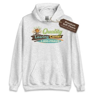 May include: A light gray hoodie with a front pocket and a hood. The front of the hoodie features a graphic with the words "Quality Learing Center" and the phone number "1-800-FRAUD". A tag reads "Premium Soft Fleece".