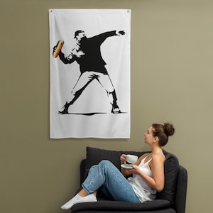 May include: White wall flag featuring a black and white stencil art design of a figure throwing a hot dog. The figure is wearing a hat and jacket. The hot dog is yellow and red.
