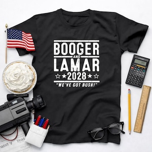May include: Black t-shirt with the text "BOOGER AND LAMAR 2028 WE'VE GOT BUSH!" in white. An American flag, a pie, a calculator, a pencil, a ruler, a film camera, a pack of pens, and glasses are arranged around the shirt.