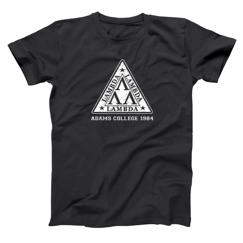 Pu&ograve; includere: T-shirt nero con un design triangolare bianco. Il design include la lettera greca Lambda, stelle e la scritta "ADAMS COLLEGE 1984". Lo stile generale evoca un design universitario vintage.