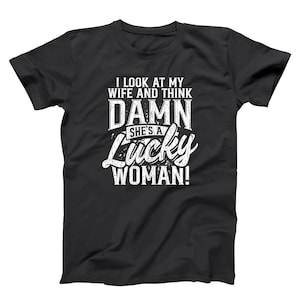 May include: Black t-shirt with white text that reads "I LOOK AT MY WIFE AND THINK DAMN SHE'S A Lucky WOMAN!" The text is in a bold, stylized font. The shirt is a crew neck style.