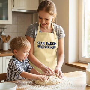 Op de afbeelding: Een lichtcr&egrave;mekleurig schort met de tekst STAR BAKER in dikke blauwe letters en een decoratief blauw krulmotief eronder. Het schort is voorzien van een nekband en taillebanden voor volledige bedekking van de romp tijdens het bakken.