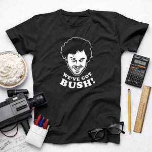 May include: Black t-shirt with a white graphic of a man's face and the text "WE'VE GOT BUSH!". The shirt is surrounded by a pie, a calculator, a pencil, a ruler, a video camera, glasses, and pens.