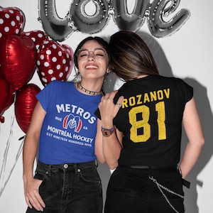 May include: Two people wearing t-shirts, one blue and one black, stand in front of a white wall with silver balloons spelling "Love" and red heart-shaped balloons. The blue shirt says "Metros Montréal Hockey". The black shirt has "Rozanov 81".