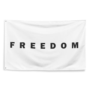 May include: White rectangular flag with the word "FREEDOM" printed in large, bold, black capital letters. The flag has grommets in the corners for hanging. The flag is made of a soft, slightly textured fabric.