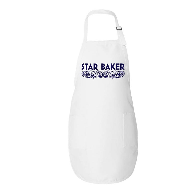 Op de afbeelding: Een wit schort met de tekst STAR BAKER in donkerblauwe hoofdletters, geplaatst boven een decoratief donkerblauw krulmotief. Het schort heeft een nekband met een verstelbare metalen schuif en een enkele zak aan de rechterkant.