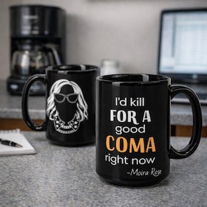 Może przedstawiać: Biały ceramiczny kubek z czarnym uchwytem i napisem "But First, Coffee" pogrubioną, czarną czcionką. Kubek jest ustawiony na rozmytym tle, podkreślając jego czysty design.