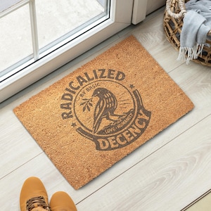 May include: A rectangular doormat made of natural fibers. The mat features a circular emblem with a bird holding a branch, surrounded by the words "RADICALIZED BY BASIC DECENCY LOVE HUMANITY". The mat is placed near a door.