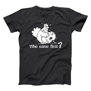 May include: Black t-shirt with a white graphic of a chicken looking at a cracked egg. The text "Who came first?" is below the graphic. The design is a humorous take on the classic chicken or the egg riddle.