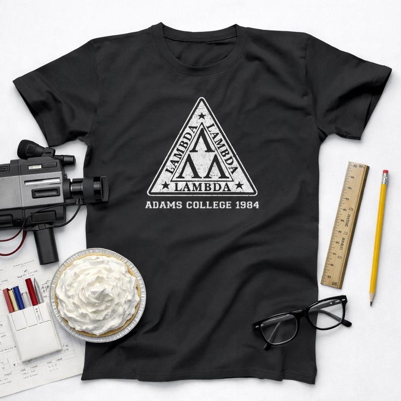 Pu&ograve; includere: T-shirt nero con logo triangolare Lambda bianco e la scritta "ADAMS COLLEGE 1984". La maglietta &egrave; mostrata con una torta, una telecamera vintage, occhiali, un righello e una matita.