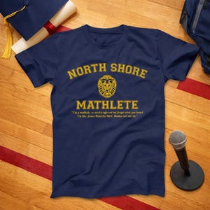 May include: Navy blue t-shirt with gold text that reads "NORTH SHORE MATHLETE". A lion graphic is centered above the word "MATHLETE". The quote "I'm a mathlete, so nerd is inferred but forget what you heard. I'm like James Bond the third. Shaken not stirred." is below.