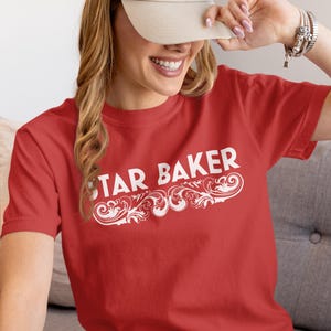 May include: A red t-shirt with the text "STAR BAKER" in white script and decorative flourishes. The person in the image is wearing a beige baseball cap.