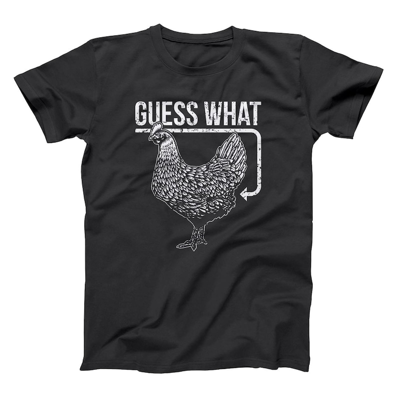 May include: Black t-shirt with a white graphic of a chicken and the text "GUESS WHAT". The chicken illustration is detailed, with a textured appearance. The design includes an arrow pointing towards the chicken, adding visual interest. A casual, everyday wear tee.