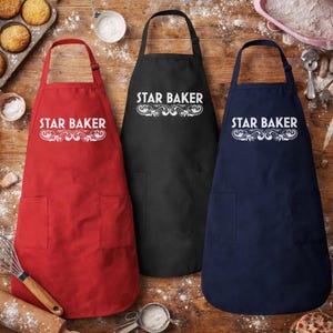 May include: Three aprons in red, black, and navy blue, each with a pocket and the text "STAR BAKER" in white script. The aprons are displayed on a wooden surface, suggesting a baking theme.