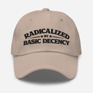 May include: A beige baseball cap featuring the black text "RADICALIZED BY BASIC DECENCY". The text is arched and centered on the front. The cap has a curved peak and a button on the crown.